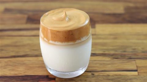 A clear glass of dalgona coffee with a layer of whipped coffee foam on top of milk. The background surface is a wooden table.