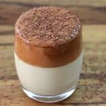A clear glass filled with a two-layered drink sits on a wooden surface. The bottom layer is white and creamy, while the thick, frothy top layer is a rich brown. Chocolate shavings are sprinkled on top and scattered around the glass.