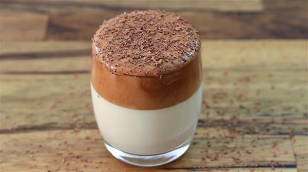 A clear glass filled with a two-layered drink sits on a wooden surface. The bottom layer is white and creamy, while the thick, frothy top layer is a rich brown. Chocolate shavings are sprinkled on top and scattered around the glass.