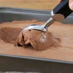 A hand holding a black-handled ice cream scooper is scooping out a portion of chocolate ice cream from a rectangular metal container on a wooden surface.