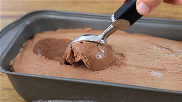 A hand holding a black-handled ice cream scooper is scooping out a portion of chocolate ice cream from a rectangular metal container on a wooden surface.