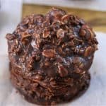 A stack of three no-bake chocolate oatmeal cookies sits on a piece of parchment paper on a wooden surface. The cookies are irregularly shaped and have a glossy, chocolaty appearance, studded with oats and small bits.