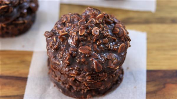 A stack of three no-bake chocolate oatmeal cookies sits on a piece of parchment paper on a wooden surface. The cookies are irregularly shaped and have a glossy, chocolaty appearance, studded with oats and small bits.