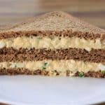 A neatly cut, triangular egg salad sandwich made with dark rye bread is stacked on a white plate, showcasing its layers filled with a creamy egg mixture and garnished with green herbs. The background features a blurred wooden surface.