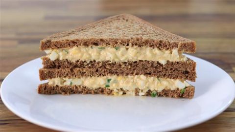 A neatly cut, triangular egg salad sandwich made with dark rye bread is stacked on a white plate, showcasing its layers filled with a creamy egg mixture and garnished with green herbs. The background features a blurred wooden surface.