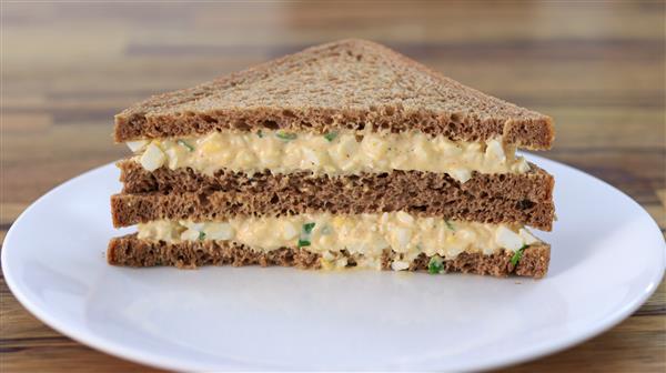 A neatly cut, triangular egg salad sandwich made with dark rye bread is stacked on a white plate, showcasing its layers filled with a creamy egg mixture and garnished with green herbs. The background features a blurred wooden surface.