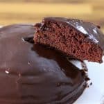 A chocolate cake with glossy frosting sits on a white plate. A slice of the cake is being lifted with a metal spatula, revealing the moist, rich chocolate interior. The background is a wooden surface.