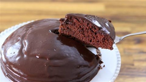 A chocolate cake with glossy frosting sits on a white plate. A slice of the cake is being lifted with a metal spatula, revealing the moist, rich chocolate interior. The background is a wooden surface.