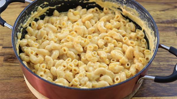 A close-up of a pan filled with creamy macaroni and cheese. The dish is composed of elbow macaroni pasta mixed with a rich, melted cheese sauce. The pan has black handles and is placed on a wooden surface.