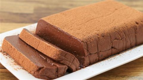 A rectangular chocolate terrine sits on a white plate. The dessert has a smooth texture and is dusted with cocoa powder on top. Two thick slices have been cut and are placed in front of the main terrine, showing the rich, chocolatey interior.