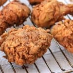 Several pieces of golden-brown, crispy fried chicken rest on a wire rack, allowing excess oil to drain onto the white paper towels beneath. The chicken pieces vary in size, including drumsticks, breasts, and thighs.