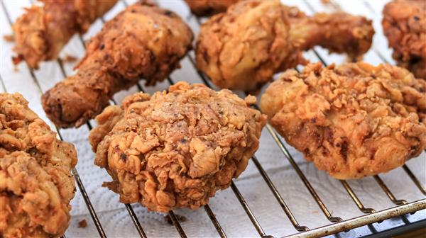 Several pieces of golden-brown, crispy fried chicken rest on a wire rack, allowing excess oil to drain onto the white paper towels beneath. The chicken pieces vary in size, including drumsticks, breasts, and thighs.