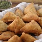 A woven basket lined with white paper holds several golden-brown, triangular samosas. In the background, two small bowls of dipping sauces are visible, one green and one red. The scene is set on a wooden surface.