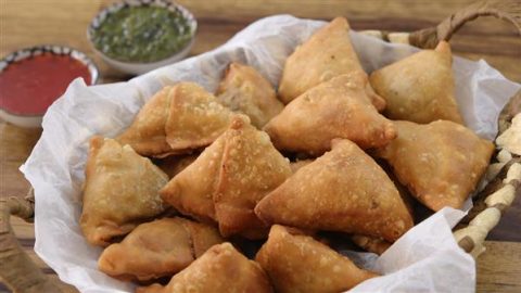 A woven basket lined with white paper holds several golden-brown, triangular samosas. In the background, two small bowls of dipping sauces are visible, one green and one red. The scene is set on a wooden surface.