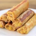A white plate with a stack of fried, crispy banana egg rolls (turon). The rolls are golden-brown and appear to have a sweet filling. The background is slightly out of focus, highlighting the appetizing appearance of the food.