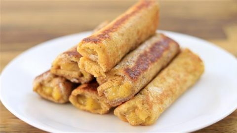 A white plate with a stack of fried, crispy banana egg rolls (turon). The rolls are golden-brown and appear to have a sweet filling. The background is slightly out of focus, highlighting the appetizing appearance of the food.