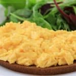 Close-up of a plate with scrambled eggs on top of a slice of brown toast, accompanied by a side of mixed leafy green salad. The dish is presented on a white plate.