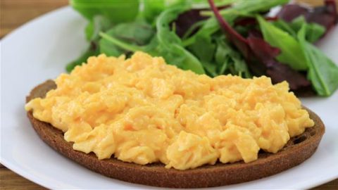 Close-up of a plate with scrambled eggs on top of a slice of brown toast, accompanied by a side of mixed leafy green salad. The dish is presented on a white plate.