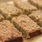 A close-up image of eight homemade granola bars on a wooden surface. The bars have a golden-brown, textured appearance, with visible oats and various nuts and seeds, arranged in two rows.