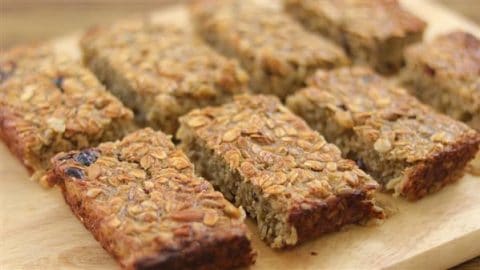 A close-up image of eight homemade granola bars on a wooden surface. The bars have a golden-brown, textured appearance, with visible oats and various nuts and seeds, arranged in two rows.