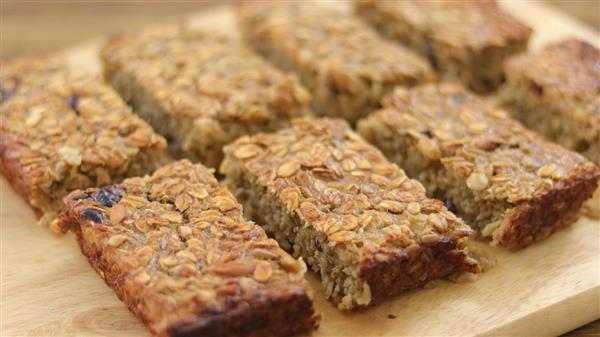 A close-up image of eight homemade granola bars on a wooden surface. The bars have a golden-brown, textured appearance, with visible oats and various nuts and seeds, arranged in two rows.