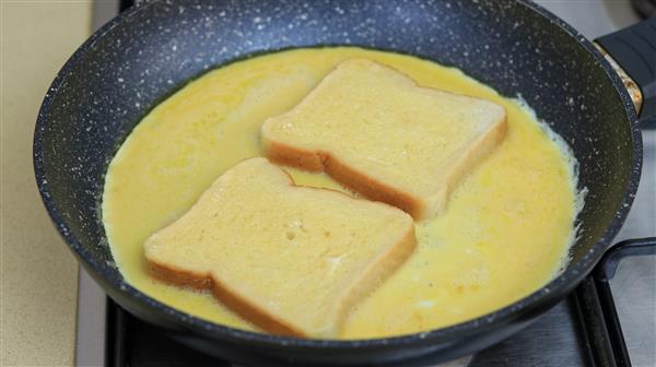 Slices of bread are soaking in beaten eggs in a nonstick frying pan on a stovetop.