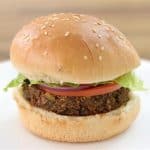 A vegan burger with a sesame seed bun on a white plate. The burger consists of a patty made from plant-based ingredients, topped with a slice of tomato, red onion, and a piece of lettuce. The background is a blurred wooden surface.