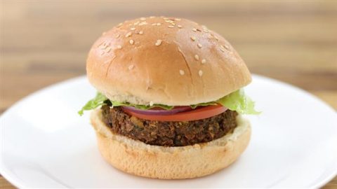 A vegan burger with a sesame seed bun on a white plate. The burger consists of a patty made from plant-based ingredients, topped with a slice of tomato, red onion, and a piece of lettuce. The background is a blurred wooden surface.