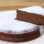 A slice of rich, fudgy chocolate cake dusted with powdered sugar is being lifted from a larger cake on a white plate. The wooden background highlights the cake's dark, moist interior.