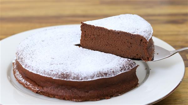 A slice of rich, fudgy chocolate cake dusted with powdered sugar is being lifted from a larger cake on a white plate. The wooden background highlights the cake's dark, moist interior.