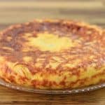 A golden-brown Spanish tortilla, or omelette, sits on a clear glass plate. The texture appears fluffy and slightly crispy on the outside. The background features a wooden surface.