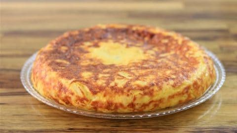 A golden-brown Spanish tortilla, or omelette, sits on a clear glass plate. The texture appears fluffy and slightly crispy on the outside. The background features a wooden surface.