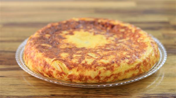 A golden-brown Spanish tortilla, or omelette, sits on a clear glass plate. The texture appears fluffy and slightly crispy on the outside. The background features a wooden surface.