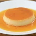 A creamy flan dessert topped with caramel sauce is served on a white plate. The caramel sauce creates a shiny, golden-brown pool around the flan, enhancing its appetizing appearance. The plate rests on a wooden surface.