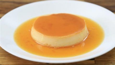 A creamy flan dessert topped with caramel sauce is served on a white plate. The caramel sauce creates a shiny, golden-brown pool around the flan, enhancing its appetizing appearance. The plate rests on a wooden surface.