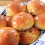 A basket lined with a blue and white checkered cloth holds several freshly baked, golden brown buns with a slight shine on their surface. The buns appear soft and fluffy, lightly browned to perfection.