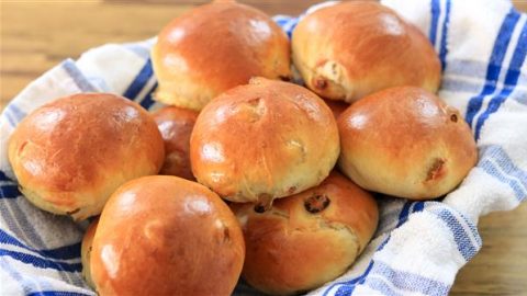 A basket lined with a blue and white checkered cloth holds several freshly baked, golden brown buns with a slight shine on their surface. The buns appear soft and fluffy, lightly browned to perfection.