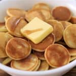 A white bowl filled with small round pancakes topped with a square pat of butter and drizzled with syrup. The bowl is placed on a wooden surface.