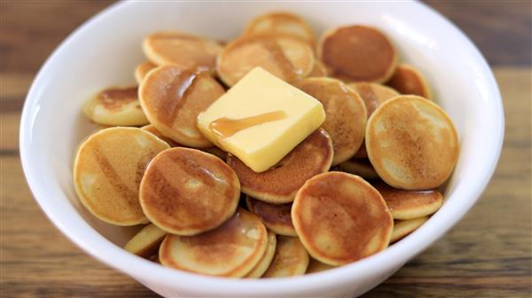 A white bowl filled with small round pancakes topped with a square pat of butter and drizzled with syrup. The bowl is placed on a wooden surface.