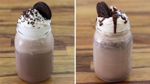 Two mason jar milkshakes sit side by side on a wooden table. Each is topped with whipped cream, an Oreo cookie, and chocolate crumbs. The milkshake on the right also has a drizzle of chocolate syrup over the whipped cream.