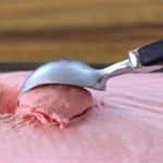 A hand using a metal ice cream scoop to scoop pink ice cream from a rectangular container on a wooden surface. The ice cream appears to be soft and creamy.