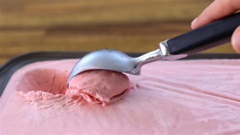 A hand using a metal ice cream scoop to scoop pink ice cream from a rectangular container on a wooden surface. The ice cream appears to be soft and creamy.