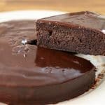 A rich, glossy chocolate cake with a smooth chocolate ganache topping sits on a white plate. A slice is being lifted from the cake, revealing the moist and dense interior. The background is a wooden surface.