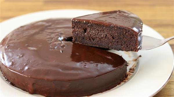 A rich, glossy chocolate cake with a smooth chocolate ganache topping sits on a white plate. A slice is being lifted from the cake, revealing the moist and dense interior. The background is a wooden surface.