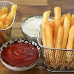 Two metal baskets filled with golden, crispy French fries are placed on a wooden table. Between them, there are two small bowls, one filled with ketchup and the other with a creamy white sauce.