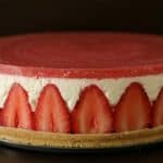 A beautifully crafted strawberry cheesecake with a golden biscuit crust, a creamy white filling, and a topping of vibrant strawberry gel. Sliced strawberries line the outer edge, adding a decorative and mouth-watering touch.