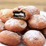 A plate of deep-fried Oreo cookies covered in powdered sugar. One cookie is split open, showing the black and white Oreo filling inside. The treats are golden brown and look crispy on the outside, resting on a white plate on a wooden surface.