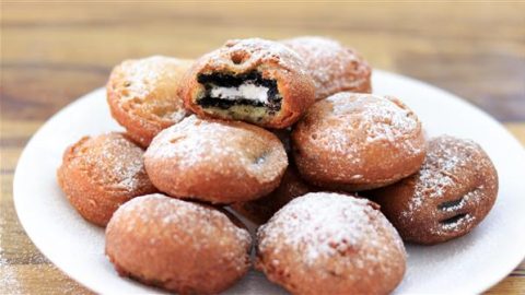 A plate of deep-fried Oreo cookies covered in powdered sugar. One cookie is split open, showing the black and white Oreo filling inside. The treats are golden brown and look crispy on the outside, resting on a white plate on a wooden surface.