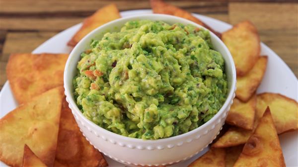 A white bowl filled with creamy guacamole sits on a plate surrounded by crispy, golden-brown tortilla chips. The guacamole is vibrant green with visible chunks of ingredients, such as tomato and herbs. The background consists of a wooden surface.