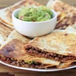 A plate of golden-brown quesadillas, filled with seasoned ground beef, sits on a wooden surface. The quesadillas are neatly arranged around a small white bowl of guacamole placed in the center of the plate.